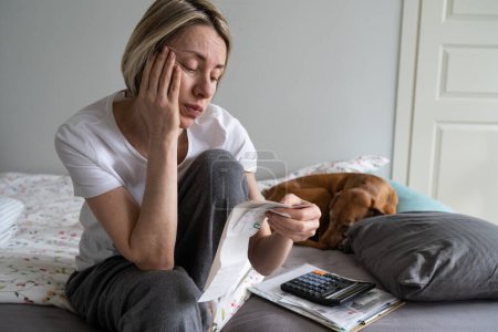 Upset middle aged woman with pay bills and calculator sits on bed with dog in home m interior. Perplexed mature female with financial problems stressed when sees too much rent or electricity costs