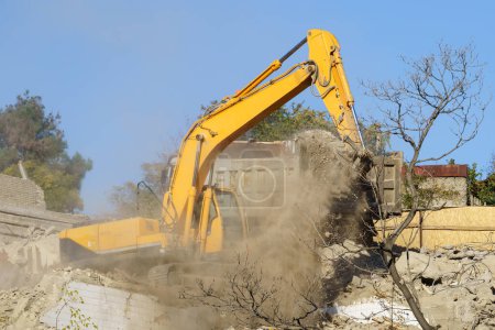 Photo for Yellow crawler excavator demolishes dilapidated real estate for future construction of new modern house. Clouds of dust around special construction equipment cleaning up rubble of collapsed building - Royalty Free Image