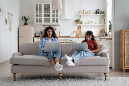 Photo for Modern introverted woman and teenage girl sits on sofa at home with laptops on laps. Unsociable Caucasian mother and child in casual clothes are addicted to gadgets and excessive use of internet - Royalty Free Image