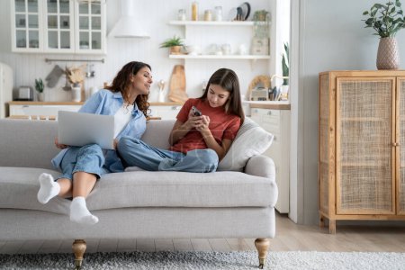Independent everyday teen girl keeps secrets from mom by hiding phone screen and avoiding surveillance. Smiling young woman with laptop on lap sits on sofa trying to make friends with daughter