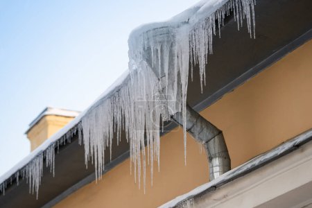 Grandes carámbanos congelados que cuelgan peligrosamente del borde del edificio en el frío día de invierno, formación de hielo peligroso en el techo de la casa de metal durante el soleado brillante, pero el clima subcongelante afuera. Prevención de presas de hielo