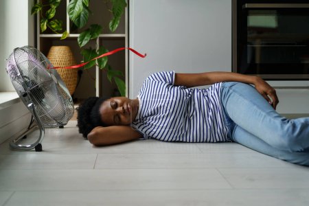 Foto de Mujer africana sobrecalentada acostada en el suelo durmiendo con ventilador eléctrico encendido, disfrutando del aire fresco, hembra negra agotada descansando en casa durante el clima extremadamente caliente. Mantenerse fresco sin aire acondicionado - Imagen libre de derechos
