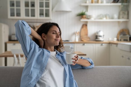 Happy relaxed woman enjoying lazy day at home, holding cup of tea and stretching body with closed eyes, resting on sofa in cozy apartment, taking break from daily routine, drinking morning coffee