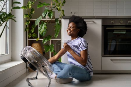 Téléchargez les photos : Jeune femme heureuse satisfaite afro-américaine assise sur le sol dans la cuisine en face de refroidisseur d'air, fille noire utilisant ventilateur pour se refroidir à la maison. Heureuse femme heureuse se refroidissant avec ventilateur - en image libre de droit