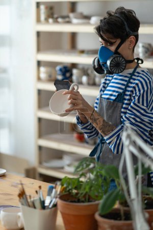 Téléchargez les photos : Young female artisan concentrated on modelling mug from clay and polishes the finished product in a respirator in creative studio. Woman ceramic business owner making craft for sale - en image libre de droit
