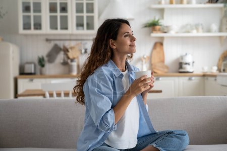 Pleased smiling woman resting on sofa with cup of tea enjoying alone time at home, looking aside with dreamy face expression. Positive female starting day with morning coffee. Free time for parent