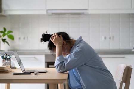 Téléchargez les photos : Stressé femme freelance frustré assis devant un ordinateur portable dans la cuisine, tenant la tête dans les mains, aux prises avec la tâche tout en travaillant à distance. Travail à domicile, côté émotionnel du freelance - en image libre de droit