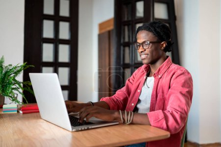 Téléchargez les photos : Heureux jeune Africain satisfait freelance gars assis au bureau à la maison en utilisant un ordinateur portable, regardant l'écran d'ordinateur et souriant, profiter de travailler à distance. Travail indépendant, travail indépendant et santé mentale - en image libre de droit
