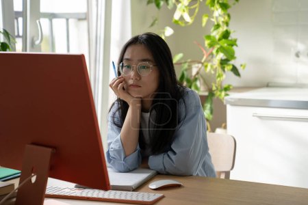 Photo for Young serious Asian girl student watching webinar video course on desktop computer while studying at home, staying focused during online learning, gaining knowledge from internet. Self-education - Royalty Free Image