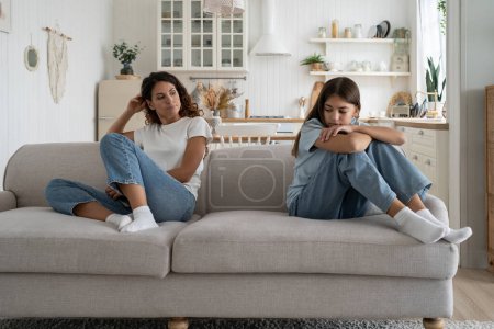 Upset teenage daughter and young woman mother sitting separately in silence on sofa at home, feeling frustrated after quarrel. Sad parent mom having misunderstandings with troubled teen girl child