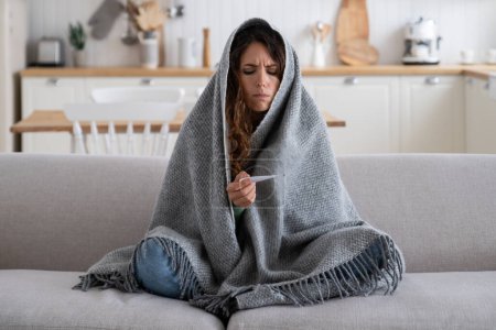 Téléchargez les photos : Sick unhappy woman frustrated looking at thermometer having cold during winter walk or work on frost. Diseased sad girl wrapped in blanket has chills and stress sits on sofa in unheated apartment - en image libre de droit
