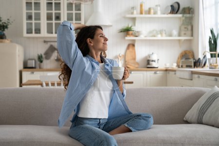 Happy dreamy young woman wearing casual clothes resting with cup of tea at cozy home, smiling female sitting on couch enjoying favorite hot drink, sipping it slowly to relax. Hygge and slow life