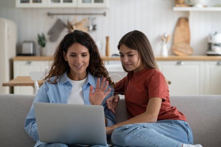 Foto de Smiling positive woman nanny and school age girl making video call in laptop contacting parents teenager. Kind caring mother waving hand greeting Internet interlocutor sits on couch with daughter - Imagen libre de derechos