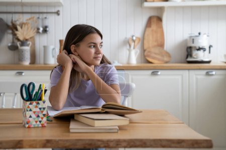 Foto de Dreamy optimistic teenage girl sits at kitchen table with books distracted from doing extracurricular work looking to side. Procrastinating youth schoolgirl cant concentrate reading textbooks - Imagen libre de derechos