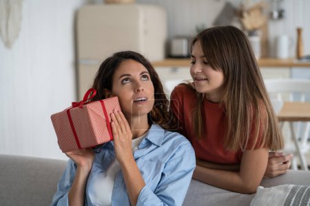 Téléchargez les photos : Happy festive family of woman and teenage girl giving mother gift box on holiday or birthday. Kind positive daughter stands behind nanny sits on sofa in living room, intrigued by unexpected president - en image libre de droit
