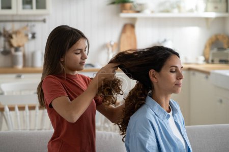 Téléchargez les photos : Family time. Pre-teen girl daughter braiding mothers long wavy hair, selective focus. Mom and child enjoying time with each other on weekend at home. Child making mommy new hairstyle - en image libre de droit