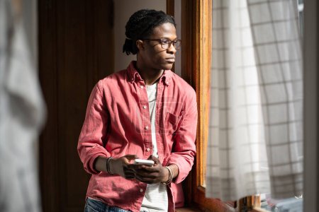Foto de Upset unhappy black guy holding smartphone looking out window, standing at home alone, frustrated depressed young African guy feeling sad after breaking up with girlfriend over phone - Imagen libre de derechos
