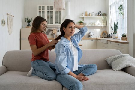 Téléchargez les photos : Teenage girl daughter braiding hair and making hairstyle for her attractive charming mother while enjoying time together at home, sitting on sofa in living room. Spending weekend with mom - en image libre de droit