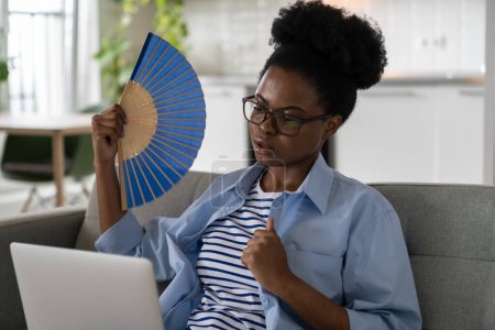 Foto de Exhausted African woman suffering from heat stroke waving blue fan and looking at laptop sits on sofa. Unhappy black girl sweat from overheating due to hot summer weather with high air temperature - Imagen libre de derechos