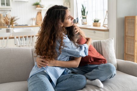 Foto de Caring loving mother comforting hugging unhappy sad teenage daughter, sitting together on sofa at home, mom supporting depressed teen girl child, parent making peace with kid. Parenting of adolescents - Imagen libre de derechos