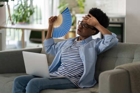Foto de Tired depressed African American millennial woman using hand fan suffering from stuffiness and non working air conditioning. Oppressed sweaty girl sits on sofa with laptop and working in hot weather - Imagen libre de derechos