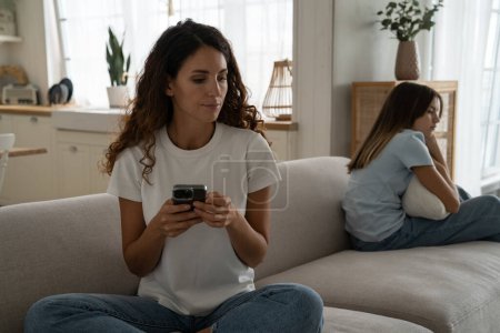 Téléchargez les photos : Unhappy sad young caucasian mother and teenage daughter sitting separately on sofa after quarrel. Upset mom holding mobile phone thinking about difficult character of adolescent child ignoring her - en image libre de droit