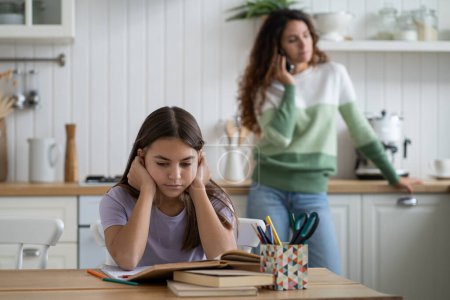 Foto de Sad tired schoolgirl overwhelmed by homework sitting at kitchen table struggling with learning while her mother standing behind and talking on phone. Parent does not care about child education - Imagen libre de derechos