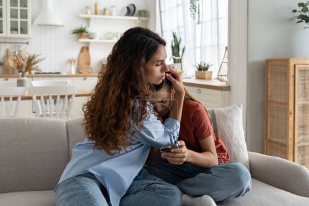 Téléchargez les photos : Upset crying teenage girl sits on sofa with phone suffering from depression after bullying. Caring kind woman hugging daughter feeling stressed because of insults in social networks or messengers - en image libre de droit