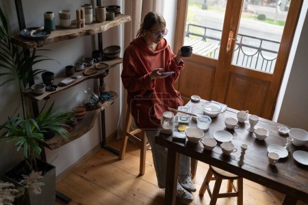 Photo for Young woman craft artist sitting at table at cozy home ceramic studio, using smartphone and drinking tea, female ceramicist promoting pottery business on social media, sell handmade ceramics online - Royalty Free Image