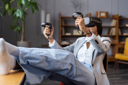 Téléchargez les photos : Casque VR pour le divertissement. Femme d'affaires portant des lunettes de réalité virtuelle utilisent joystick de jeu assis détendu avec les jambes au bureau. Travail féminin africain avec réalité augmentée ou jouer à des jeux vidéo - en image libre de droit