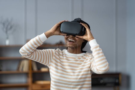 Téléchargez les photos : Opportunités en réalité virtuelle. Femme afro-américaine incroyable testant casque high-tech moderne, obtenir une expérience 3D à la maison. Joyeuse femme ethnique dans le casque VR regarder des vidéos et des films - en image libre de droit