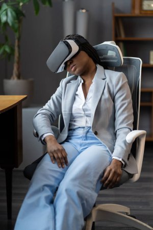 Téléchargez les photos : Fatiguée employée afro-américaine portant des lunettes de réalité virtuelle dormant dans une chaise de bureau sur le lieu de travail après avoir utilisé la technologie VR pendant trop longtemps. La cybermaladie en réalité virtuelle et augmentée - en image libre de droit