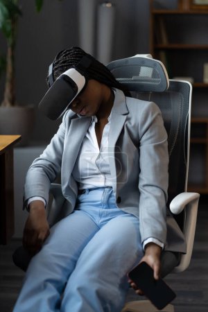 Foto de Mujer de negocios afroamericana agotada que se queda dormida usando gafas vr con teléfono inteligente en las manos. Dormir cansada en exceso de trabajo en auriculares de realidad virtual portátiles sentados en el escritorio de la oficina en el trabajo - Imagen libre de derechos