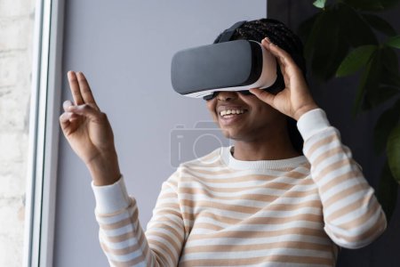 Téléchargez les photos : Jeune femme afro-américaine souriante dans les lunettes VR faisant du shopping en réalité virtuelle, explorant le magasin virtuel, ramassant et examinant des articles tout en étant assise par la fenêtre à l'intérieur. Réalité augmentée dans le commerce de détail - en image libre de droit