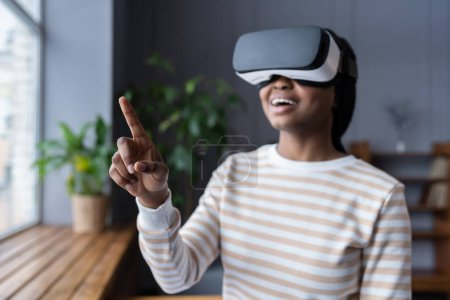 Téléchargez les photos : Black woman with open mouth wearing virtual reality glasses feeling excited and amazed while interacting with immersive digital world at cozy room decorated with green houseplants, selective focus - en image libre de droit