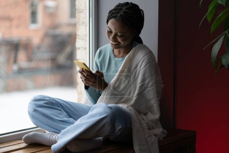 Foto de Mujer negra sonriente con teléfono celular amarillo sentado en el alféizar de la ventana en casa y hace la compra en la tienda en línea. Afro-americana joven hembra con cálido manta mensajes de texto con sus amigos en repisa por ventana - Imagen libre de derechos