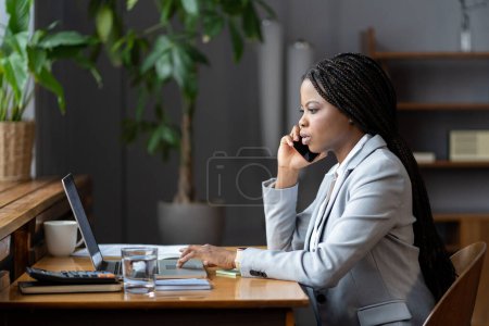 Téléchargez les photos : Effective mobile communication in workplace. Young african american woman business consultant in suit talking with client on phone and checking information on laptop, sitting in modern office interior - en image libre de droit