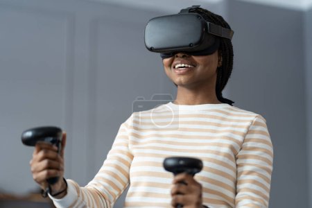 Foto de Happy young African American woman in 3D goggles playing VR games at home, smiling black girl with gaming controllers in hands imitating movements in virtual reality. Cyberspace concept. - Imagen libre de derechos