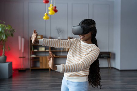 Foto de Joven mujer africana asombrada probando gafas VR tocando objetos con las manos en realidad virtual en casa. Emocionado negro millennial chica utilizando aumentada realidad en la primera vez - Imagen libre de derechos