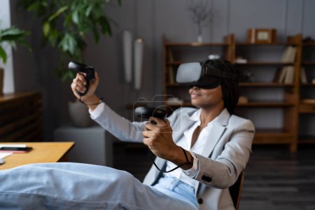 Foto de Mujer afroamericana sonriente en gafas VR sosteniendo joysticks divertirse en 3D mientras se relaja en el lugar de trabajo, enfoque selectivo. Empleada relajada jugando juegos de realidad virtual durante el descanso laboral - Imagen libre de derechos