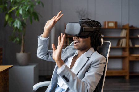 Foto de Primera experiencia de realidad virtual en el lugar de trabajo. Impresionada por la mujer negra de VR que ve videos de 360 grados en la oficina durante el descanso laboral, impresionó a la empleada africana que experimenta el mundo 3D. Enfoque selectivo - Imagen libre de derechos