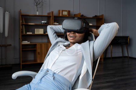Foto de Happy relaxed woman wearing VR goggles headset sit with hands behind neck at office table. Smiling businesswoman experience innovative future technology of virtual reality enjoy 3d tour or watch movie - Imagen libre de derechos