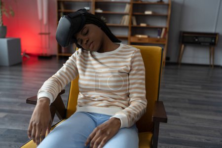 Téléchargez les photos : Fatiguée femme afro-américaine portant des lunettes de réalité virtuelle dormant dans un fauteuil à la maison après avoir utilisé la technologie VR pendant trop longtemps. Une fille qui dort. La cybermaladie en réalité virtuelle et augmentée - en image libre de droit