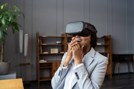Téléchargez les photos : VR headset for business experience, curious excited african american businesswoman wearing goggles. Young black female in office exploring augmented reality simulation shocked sitting with open mouth - en image libre de droit