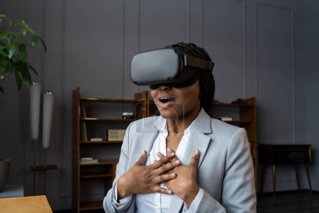 Foto de Empleada africana conmocionada emocional en auriculares VR inmersa en el mundo virtual 3D llena de emociones e impresiones, sentada en el lugar de trabajo y gritando wow durante la primera experiencia de realidad virtual - Imagen libre de derechos