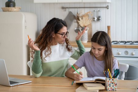 Foto de Optimistic focused teenage girl studying school curriculum reading books doing tasks under supervision of nanny. Young casual woman waving hands sits at kitchen table together with school age daughter - Imagen libre de derechos