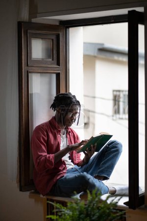 Photo for Pensive African American young man in eyeglasses reading sad book sitting on sill at home. Concentrated hipster male get knowledge from literature. Black student guy intently study difficult textbook. - Royalty Free Image