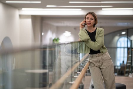 Photo for Successful self-sufficient woman talking on phone standing inside business center building calling taxi or courier. Young businesswoman leans on glass partition making call to business partners - Royalty Free Image