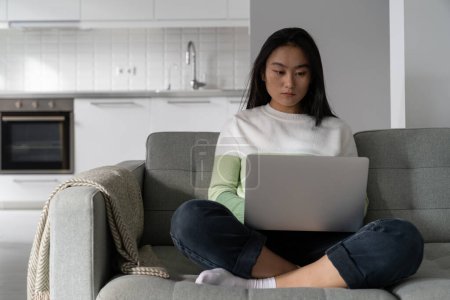 Foto de Concentrated professional korean woman sits on sofa with laptop and typing text doing remote freelance work. Self-sufficient focused Asian girl working from home located in comfortable apartment - Imagen libre de derechos