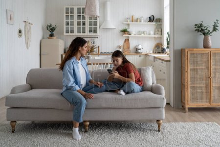 Foto de Sad teen girl holding mobile phone, experiencing cyberbullying on social media, sitting with mother on sofa at home. Mom helping daughter to cope with unrequited love. Teenage problems concept - Imagen libre de derechos